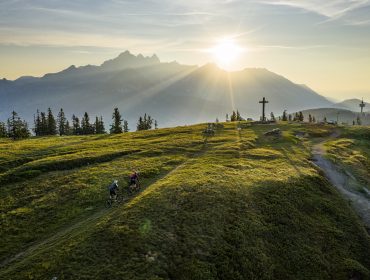 Salzburger_Sportwelt_Rossbrand_mtb_travel_2019(c)WOM_Medien-Andreas_Meyer Zwei Radfahrer fahren bei Sonnenaufgang auf einem grasbewachsenen Hügel mit Bergen, Bäumen und einem großen Kreuz im Hintergrund.