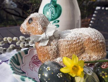 Ein lammförmiger Kuchen mit einer Schleife, bemalten Eiern und einer gelben Blume auf einem verzierten Teller.