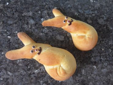 Zwei Brötchen in Hasenform mit Süßigkeitenaugen und Schokoladenmündern auf einer dunklen Arbeitsplatte.