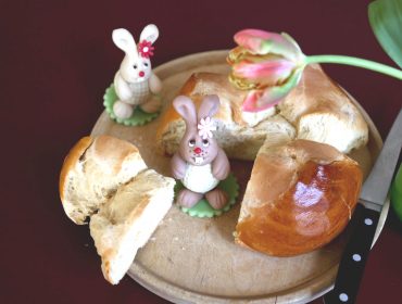 Runder Brotlaib und zwei Hasenfiguren auf einem Holzbrett, daneben eine Tulpe und ein Messer.
