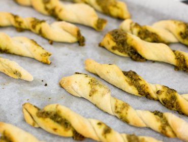 Gedrehte Brotstangen mit grüner Pestofüllung auf einem mit Pergamentpapier ausgelegten Backblech.