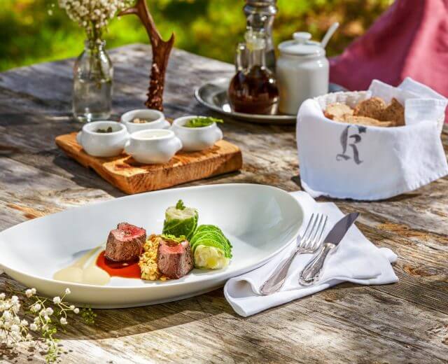 Kulinarische-Geschichten-Header Sommer Gourmet-Fleischgericht mit Saucen und Gemüse auf einem rustikalen Tisch im Freien, neben Brot, Blumen und Besteck.