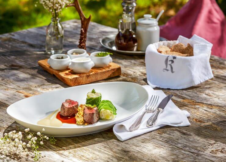 Gourmet-Fleischgericht mit Saucen und Gemüse auf einem rustikalen Tisch im Freien, neben Brot, Blumen und Besteck.