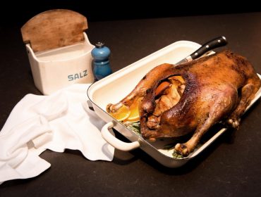 Die ganze Ente mit Kräutern und Orangenscheiben in einer Auflaufform neben einem Salzbehälter und einer blauen Pfeffermühle braten.