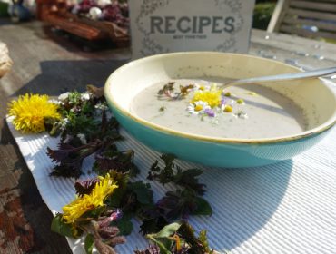 Eine Schale mit cremiger Suppe mit essbaren Blumen, neben frischen Kräutern und Löwenzahn auf einem Tisch.