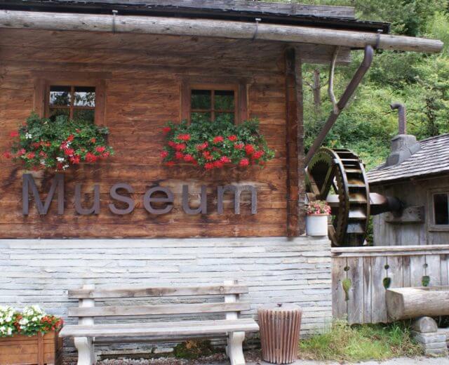 7 Muehlen Holzgebäude mit Museumsschild, Blumenkästen, einer Bank und einem Wasserrad in einer grünen Umgebung im Freien.