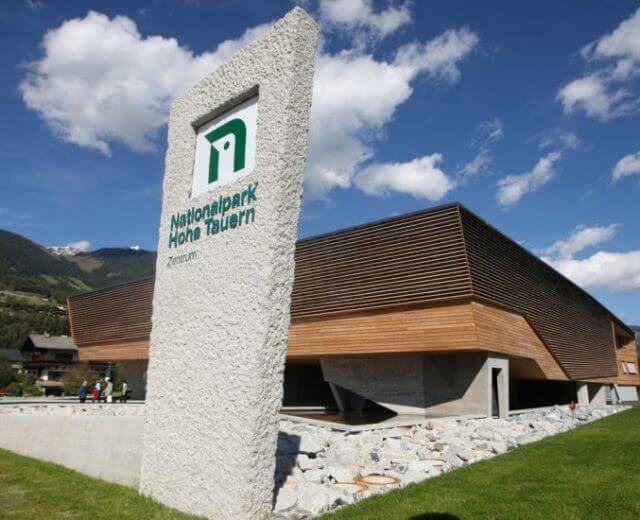 nationalparkzentrum_neu-825×550 Modernes Gebäude mit einem hohen Steinschild mit der Aufschrift Nationalpark Hohe Tauern unter einem blauen Himmel mit Wolken.