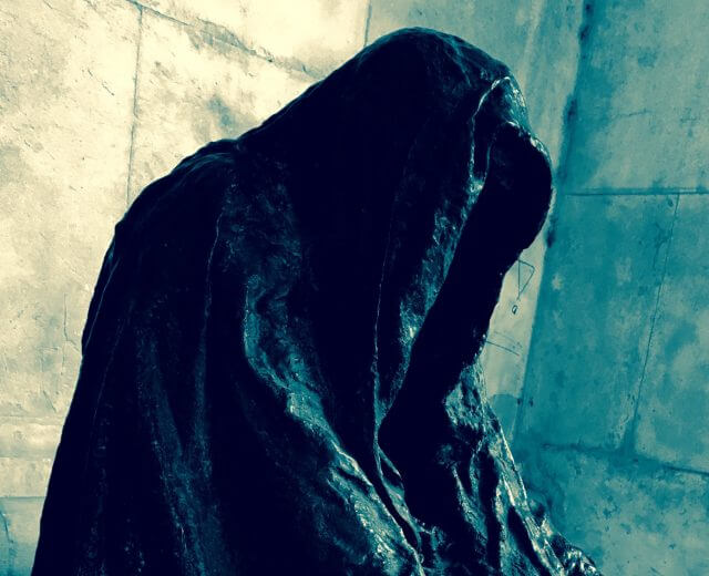 Zauberer Jackl Kapuzenlose, gesichtslose Gestalt in einem dunklen Mantel vor einer strukturierten, hellen Wand.