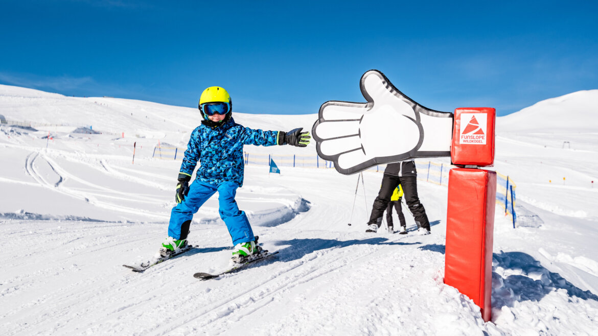 Ein Kind in blauer Skikleidung gibt einer großen Wildkogel-Schaumstoffhand auf einer verschneiten Piste unter strahlend blauem Himmel die Hand. (vergrößerte Ansicht)