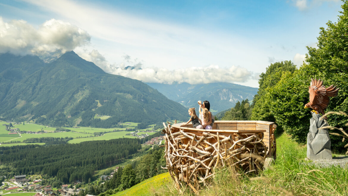 Drei Personen genießen von einer hölzernen Aussichtsplattform in Form eines Nestes oberhalb des Tales den malerischen Bergblick auf den Wildkogel. (vergrößerte Ansicht)