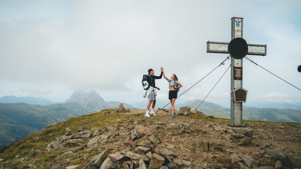 Wildkogel-Arena Zwei Wanderer geben sich neben dem großen Gipfelkreuz auf dem Wildkogel die Hand, im Hintergrund die malerische Bergkulisse. (vergrößerte Ansicht)