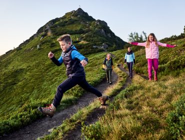Vier Kinder wandern auf einem grasbewachsenen Bergpfad; ein Junge springt über einen kleinen Graben auf dem Weg.