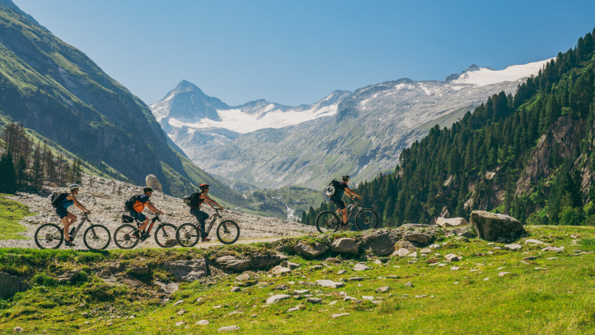 Peter Maier, Peter Maier Vier Radfahrer fahren auf der Wildkogel-Bergstrecke mit schneebedeckten Gipfeln und grünen Bäumen im Hintergrund. (vergrößerte Ansicht)