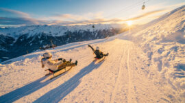 Zwei Personen rodeln bei Sonnenuntergang den verschneiten Hang des Wildkogels hinunter, mit verschneiten Gipfeln im Hintergrund.