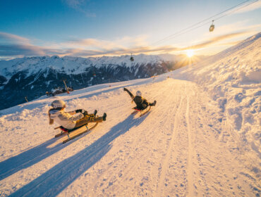Wildkogelarena_2024-3838 Zwei Personen rodeln bei Sonnenuntergang den verschneiten Hang des Wildkogels hinunter, mit verschneiten Gipfeln im Hintergrund.