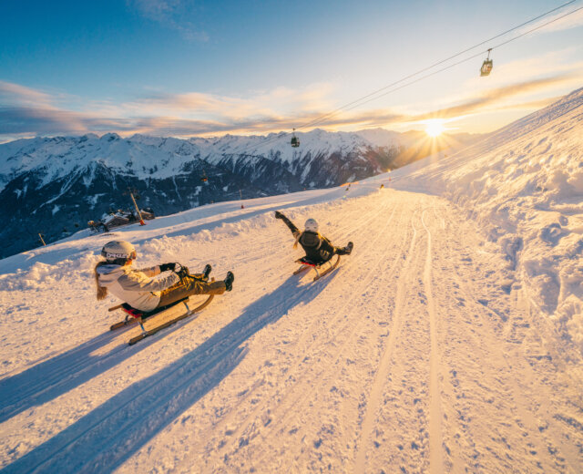 Wildkogelarena_2024-3838 Zwei Personen rodeln bei Sonnenuntergang den verschneiten Hang des Wildkogels hinunter, mit verschneiten Gipfeln im Hintergrund.
