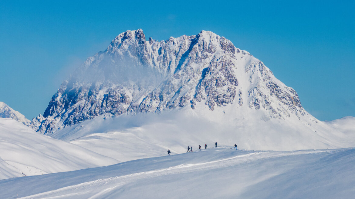 default Eine Gruppe von Skifahrern wandert über eine verschneite Wildkogel-Piste mit einem schroffen Berg im Hintergrund unter blauem Himmel. (vergrößerte Ansicht)