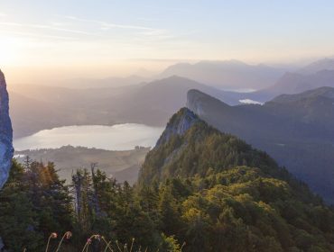 Sonnenaufgang über einer Berglandschaft mit einem See, Wald und fernen Gipfeln unter einem dunstigen Himmel.