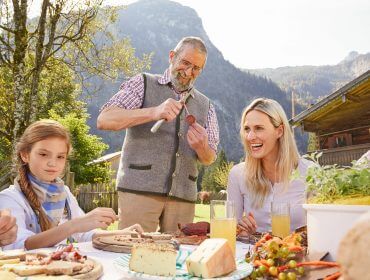 Familie beim Essen im Freien mit Bergen - So geht Herbsturlaub: Der eine schneidet eine Wurst, der andere lächelt.