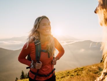 Eine lächelnde Frau in Wanderkleidung steht bei Sonnenaufgang auf einem Berg, mit Sonnenlicht und Wolken im Hintergrund.