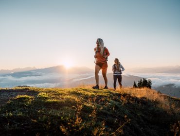 Zwei Frauen wandern bei Sonnenaufgang auf einem grasbewachsenen Hügel, mit Wolken und Bergen im Hintergrund.