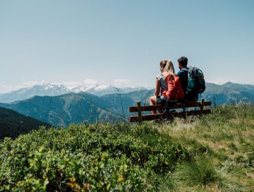 Zwei Wanderer mit Rucksäcken sitzen auf einer Bank und blicken auf grüne Berge und ferne schneebedeckte Gipfel unter einem klaren Himmel.