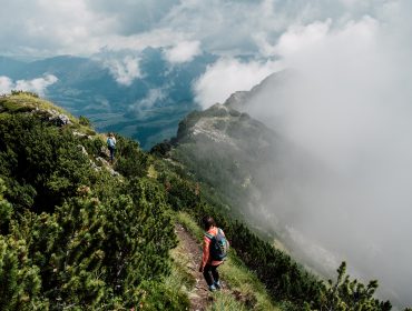 Zwei Wanderer gehen einen schmalen Bergpfad entlang, umgeben von Grün und Nebelschwaden.