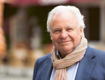 Witzigmann Lächelnder älterer Mann mit weißem Haar, der einen Schal und einen blauen Blazer trägt, steht im Freien.