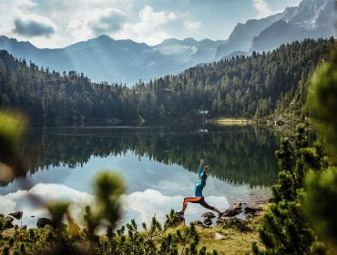 Person macht Yoga neben einem ruhigen Bergsee mit Bäumen und Bergen im Hintergrund unter einem teilweise bewölkten Himmel.