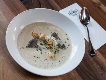 Cremesuppe mit Croutons, Kräutern und Rindfleischscheiben in einer weißen Schale auf einem Holztisch mit einem Löffel.