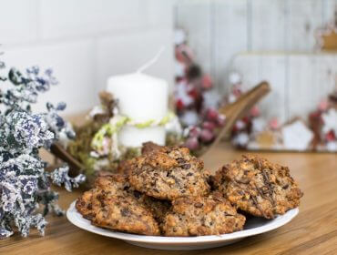 Teller mit Schokoladenkeksen auf einem Holztisch mit Weihnachtsdekoration und einer weißen Kerze im Hintergrund.