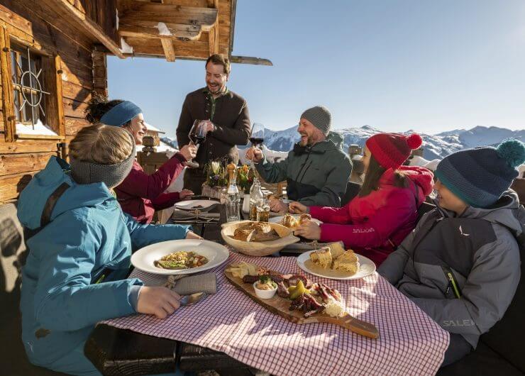 Hüttenkulinarik auf der Wolfshütte Fünf Personen in Winterkleidung genießen eine Mahlzeit vor einer Holzhütte mit verschneiten Bergen im Hintergrund.