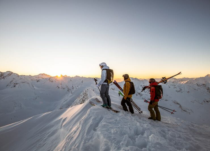 Drei Skifahrer stehen bei Sonnenaufgang auf einem verschneiten Bergrücken und blicken auf ferne, schneebedeckte Gipfel.
