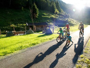 Eine Familie radelt gemeinsam auf einem sonnigen Weg in einer grünen, bergigen Landschaft mit langen Schatten hinter ihnen.
