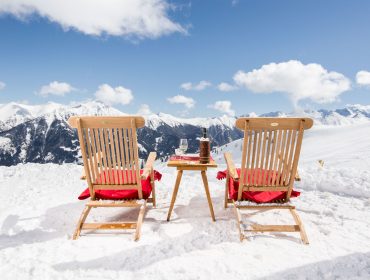 Huettenkulinarik in Gastein Zwei Holzstühle mit roten Kissen blicken auf die verschneiten Gasteiner Berge, dazwischen stehen Getränke auf einem kleinen Tisch.