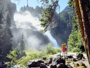 Zwei Menschen stehen auf einem felsigen Pfad im Nationalpark Hohe Tauern und bewundern einen riesigen Wasserfall im hellen Sonnenlicht.