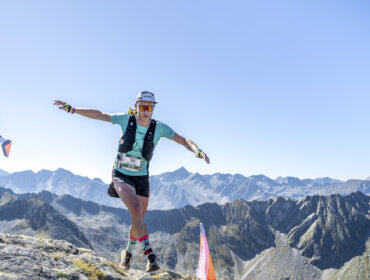 ronny_katsch_aIT23_tom_local_hero Läufer balanciert auf INFINITE TRAILS, einem felsigen Bergpfad, umrahmt von fernen Gipfeln und einem klaren blauen Himmel.