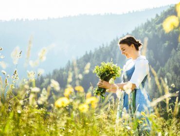 Romantik: Eine Frau auf einer Wiese mit Wildblumen, Bergen und Bäumen schafft eine romantische Atmosphäre.