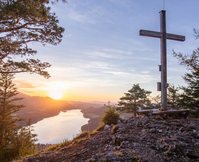 Holzkreuz auf einem Berggipfel mit Blick auf den Fuschlsee und Berge bei Sonnenuntergang, umgeben von Bäumen.