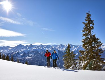 Zwei Personen in Winterausrüstung wandern auf verschneiten Pisten im Nationalpark Hohe Tauern, unter Bergen und einem strahlend blauen Himmel.