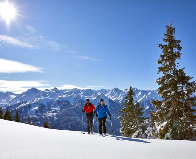 Skitour©Ferienregion Nationalpark Hohe Tauern Michael Huber Zwei Personen in Winterausrüstung wandern auf verschneiten Pisten im Nationalpark Hohe Tauern, unter Bergen und einem strahlend blauen Himmel.