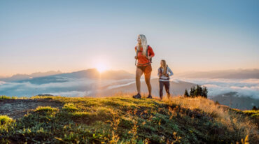 Zwei Frauen wandern bei Sonnenaufgang auf einem grünen Hügel mit Blick auf nebelverhangene Berge im Hintergrund.