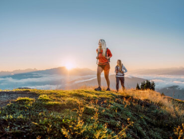 Zwei Frauen wandern bei Sonnenaufgang auf einem grünen Hügel mit Blick auf nebelverhangene Berge im Hintergrund.