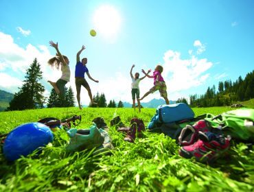 Vier Kinder springen und spielen mit einem Ball auf einer sonnigen Wiese, mit Rucksäcken und Ausrüstung im Vordergrund.