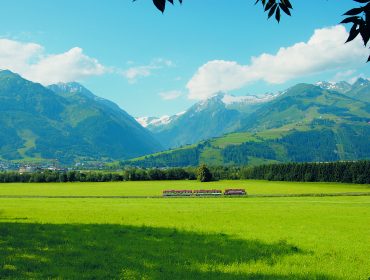 Ein roter Zug fährt durch ein grünes Tal, mit schneebedeckten Bergen im Hintergrund unter einem blauen Himmel.
