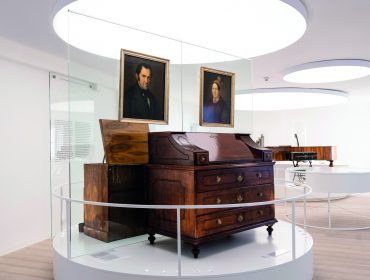 Antiker Holzschreibtisch und zwei Porträts in einer modernen, runden Museumsausstellung aus Glas.
