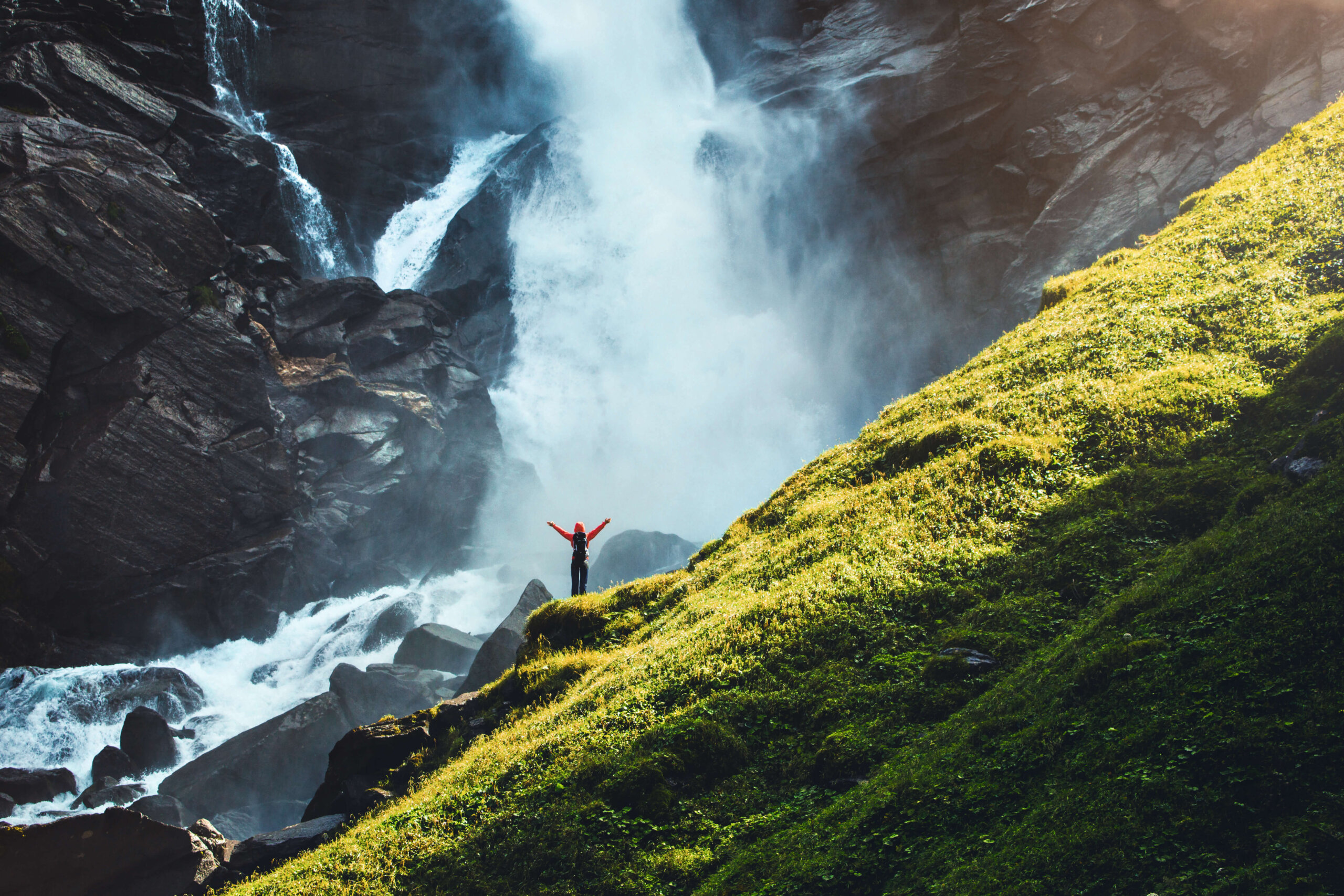 Eine Person mit erhobenen Armen steht in der Nähe eines großen Wasserfalls neben einem üppig grünen Hügel.
