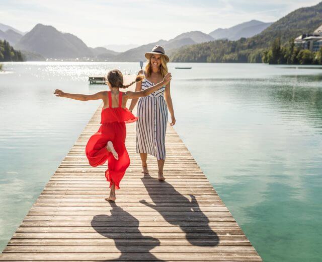 Frau und Mädchen in Sommerkleidung gehen fröhlich auf einem Holzsteg an einem klaren Bergsee spazieren.