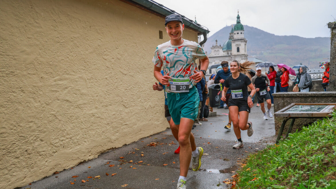 Lächelnde Läuferinnen und Läufer bei einem Berglauf auf einem nassen Weg, mit Zuschauern und einem historischen Gebäude im Hintergrund. (vergrößerte Ansicht)