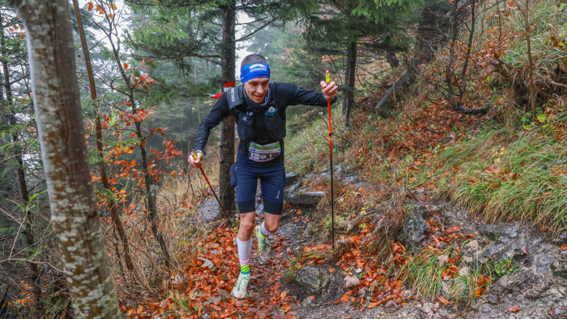 Ein Läufer genießt den Berglauf mit Trekkingstöcken auf einem schlammigen, laubbedeckten Weg durch einen Wald im Herbst. (vergrößerte Ansicht)